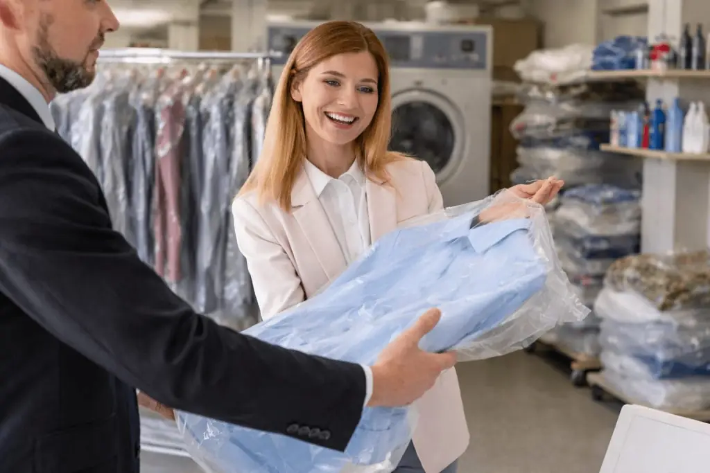 A woman smiles as she receives a freshly cleaned shirt in plastic packaging from a man at a dry cleaning near me, enjoying the convenience of laundry pickup and delivery Katy.
