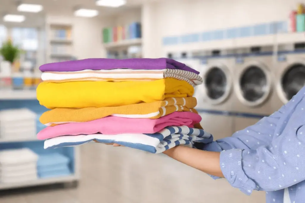 A person holds a neatly folded stack of colorful laundry inside a laundromat, with washing machines visible in the background.