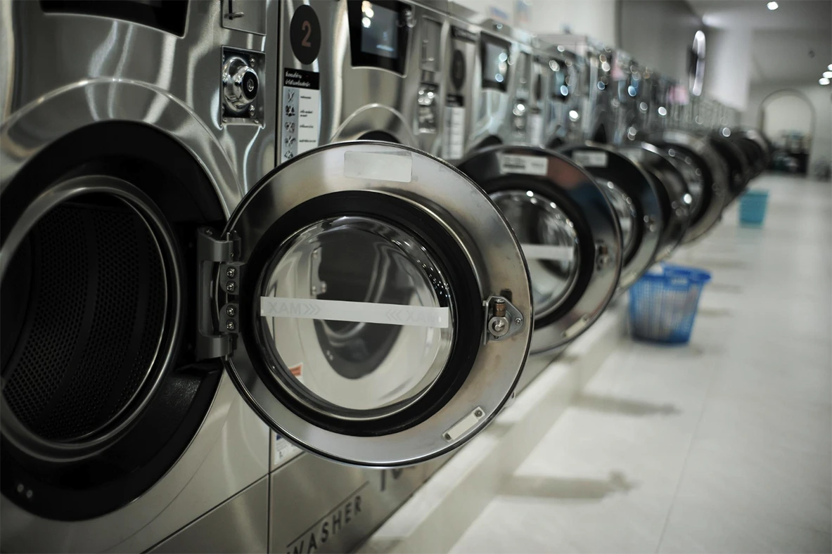 A row of stainless steel front-loading washing machines in a laundromat, with one machine door open and a blue laundry basket visible in the background.