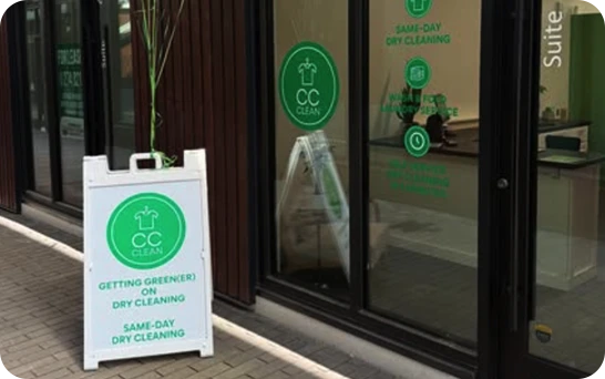 A storefront with large windows displaying "CC Clean" logos and a sidewalk sign advertising same-day dry cleaning and eco-friendly services.